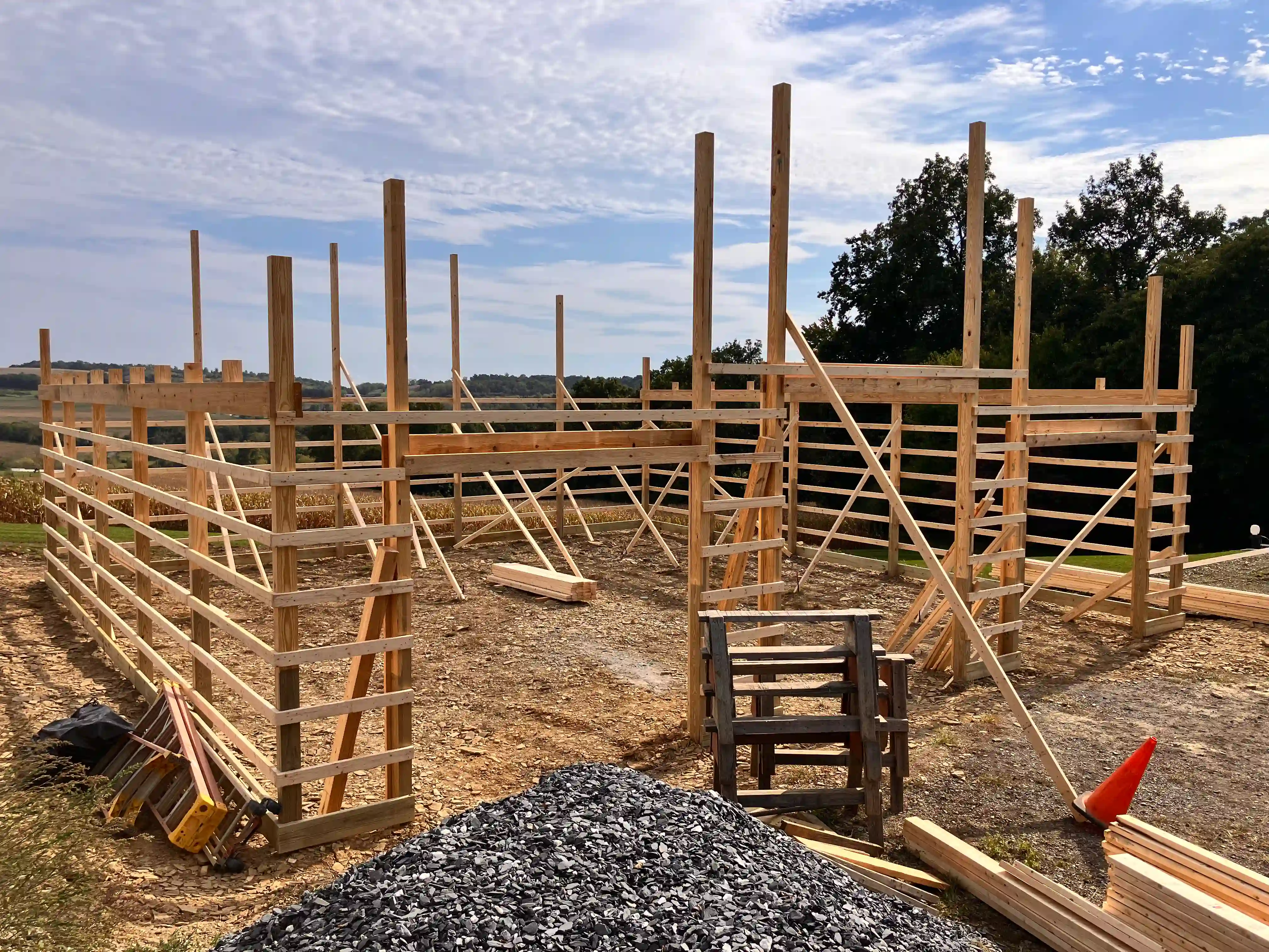 Open post-frame building under construction on a hillside, with exposed wood wall girts, diagonal bracing, and roof trusses standing against the sky